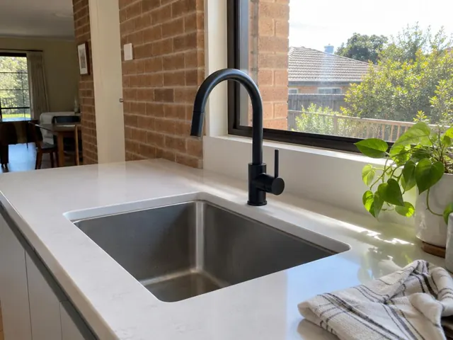 Black kitchen mixer tap above a stainless sink, with white benchtop and window in The Ponds